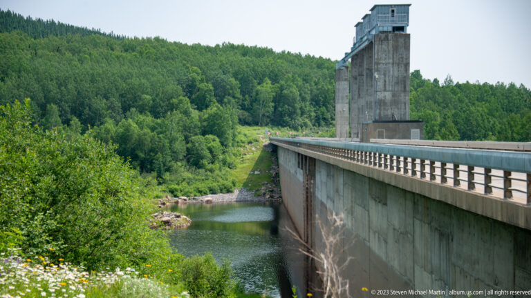 Side view of a high concrete dam wall with crest roadway and tall service tower beside forested slopes.