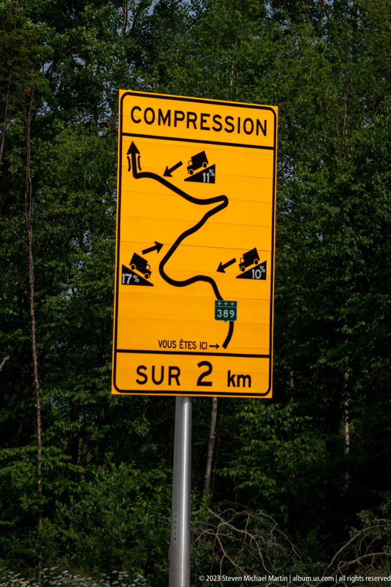 Yellow French road-warning sign labeled “COMPRESSION” showing a winding descent and vehicle guidance over two kilometres.