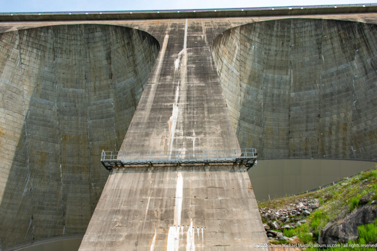 Close view of a huge concrete buttress between curved dam bays, with a maintenance platform crossing its face.