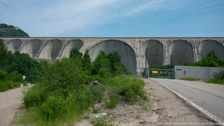 Frontal view of Daniel-Johnson Dam’s repeating concrete arches beyond a gated access road and fencing.