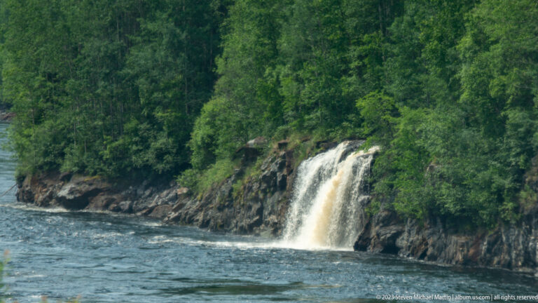 Small tan-white waterfall dropping from a forested rock bank into a broad flowing river.