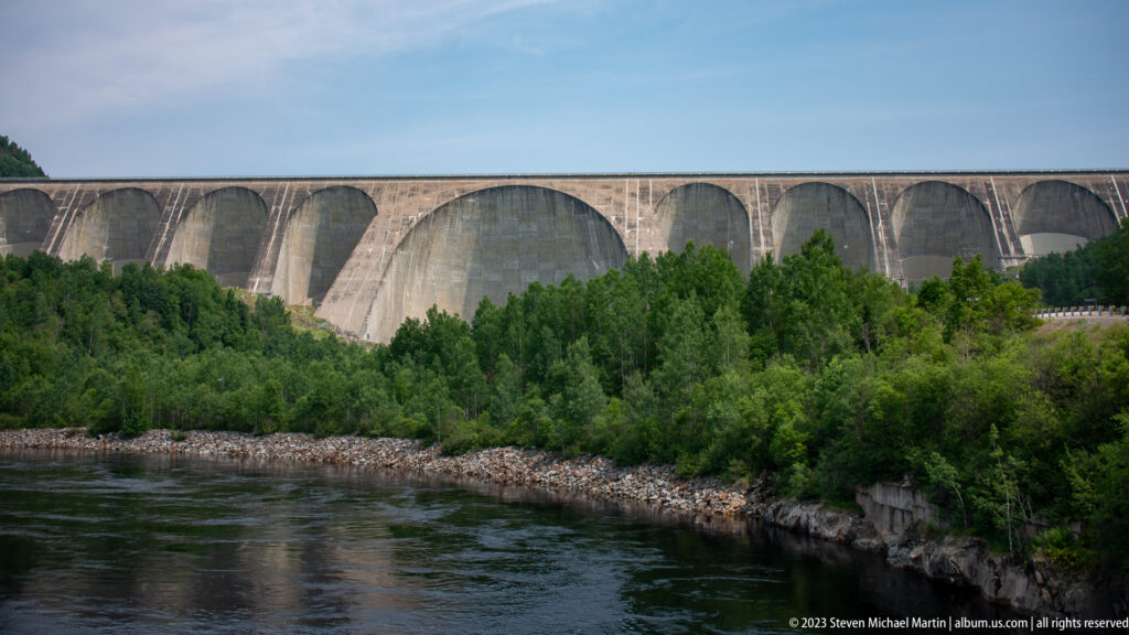 Daniel-Johnson Dam’s multiple concrete arches seen across a dark river and green shoreline.