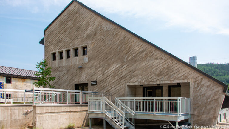Angular wood-clad building with steep roofline, narrow upper windows, and metal entrance ramps at Manic-5.