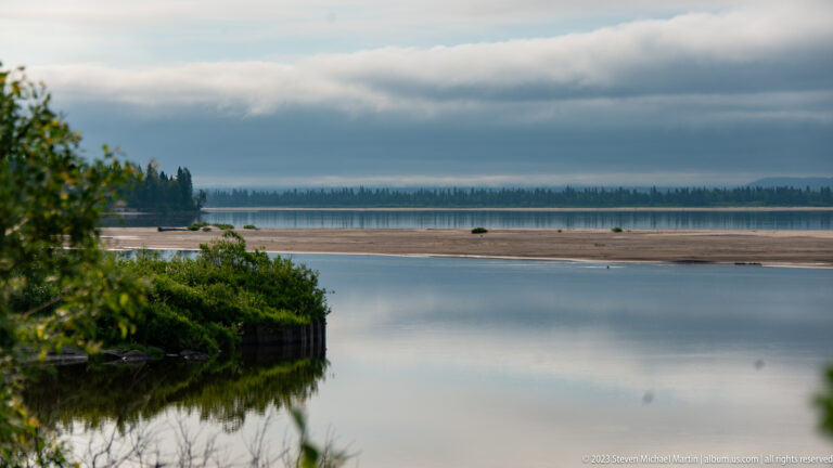 Calm channel with foreground shrubs, exposed pale flats, and a distant forested shoreline under layered clouds.