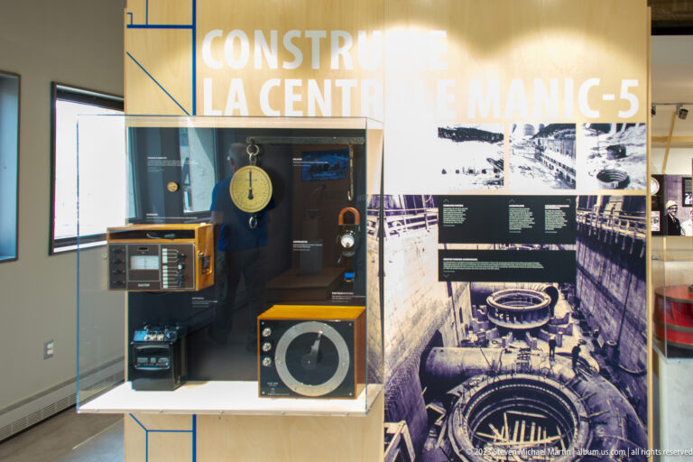 Electrical meters and control instruments displayed before a historical photograph of Manic-5 generating-station construction.