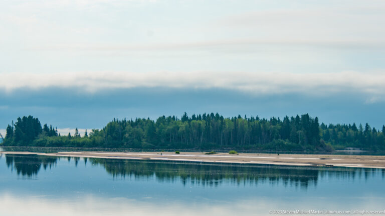 Dark conifer shoreline and pale sandbars reflected in calm water beneath soft blue-gray clouds.