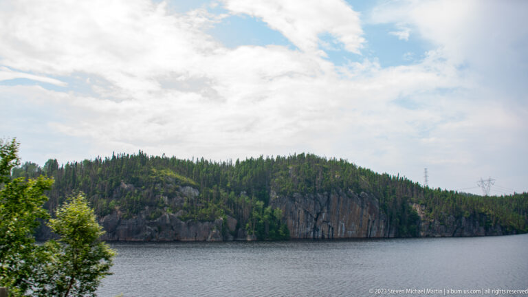 Calm reservoir water below a rocky, conifer-covered cliff and low forested hills under a bright sky.