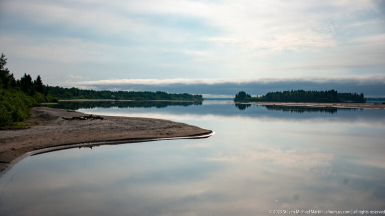 Curving sandy point extending into a calm reflective channel, with wooded islands and low shoreline in the distance.
