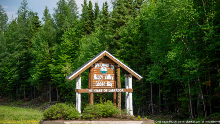 Timber welcome sign for Happy Valley-Goose Bay, “The Heart of Labrador,” standing beside a forested roadside.