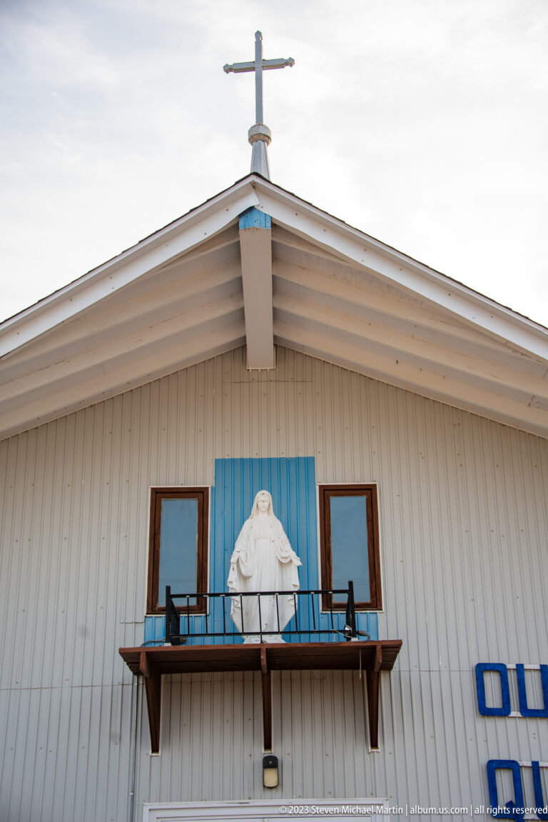 Upper façade of a pale gabled building with a roof cross, blue trim, and a white robed religious statue on a balcony.