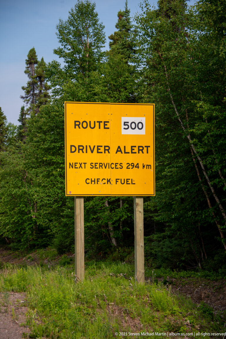 Yellow Route 500 driver-alert sign reading “Next services 294 km – Check fuel” beside dense forest.