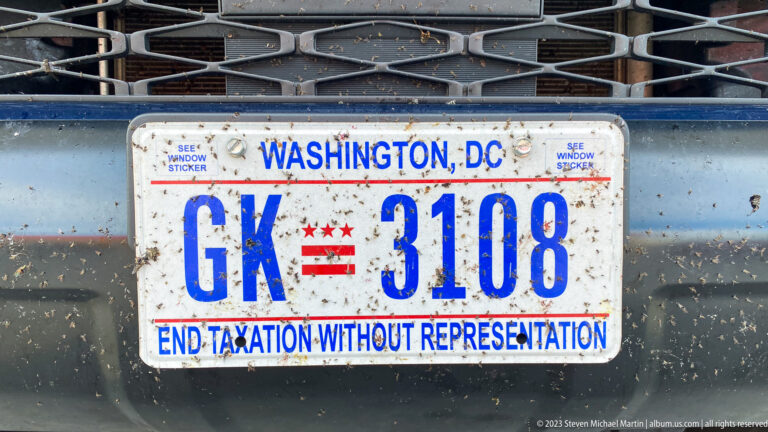Road-grimed Washington, D.C. license plate GK 3108 on the front of a vehicle, with the District slogan below.