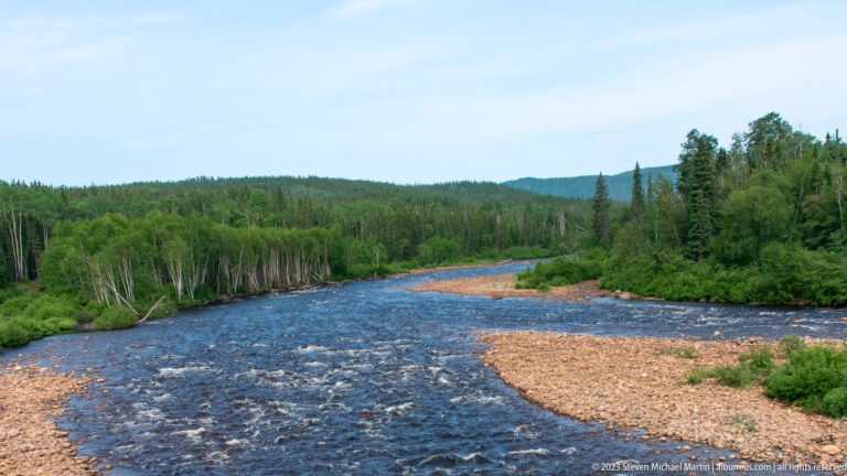 Wide fast-flowing river with low rapids, a long gravel bar, and forested hills under blue sky.