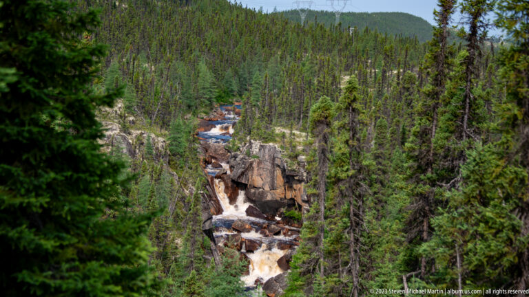 Distant cascades descending through a rocky corridor amid dense conifer forest, with a low ridge beyond.