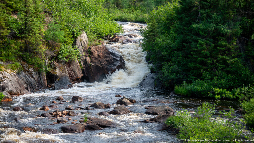 Short waterfall pouring over bedrock into a boulder-strewn river surrounded by dense green forest.