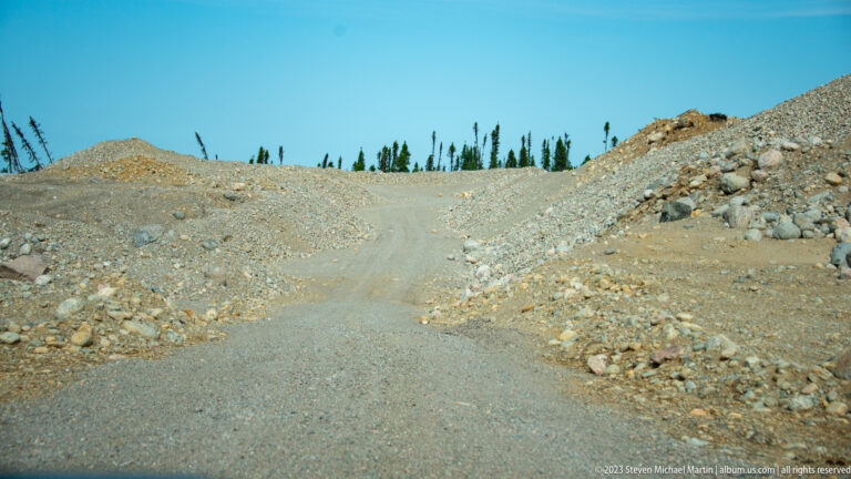 Gravel road rising between mounds of excavated rock, with sparse conifers on the horizon.