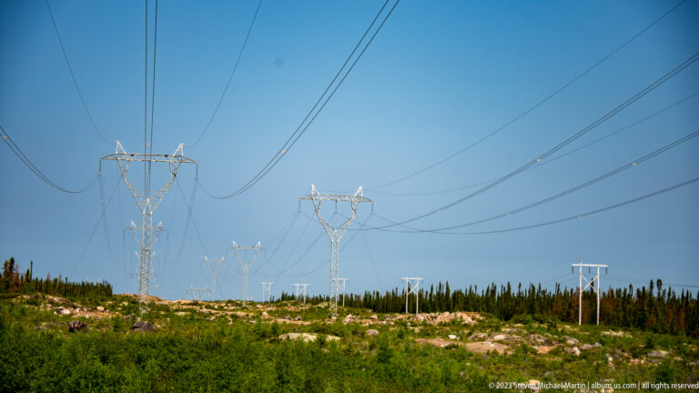 Parallel high-voltage power lines and steel transmission towers extending across low boreal terrain.