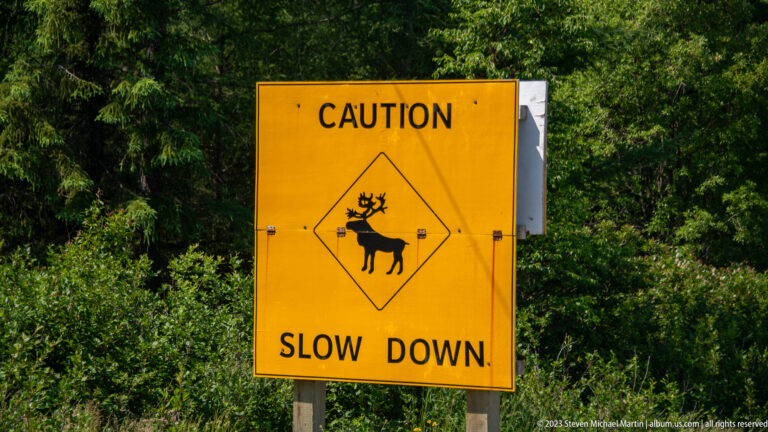 Yellow roadside caution sign reading “SLOW DOWN,” with a black silhouette of an antlered caribou-like animal.