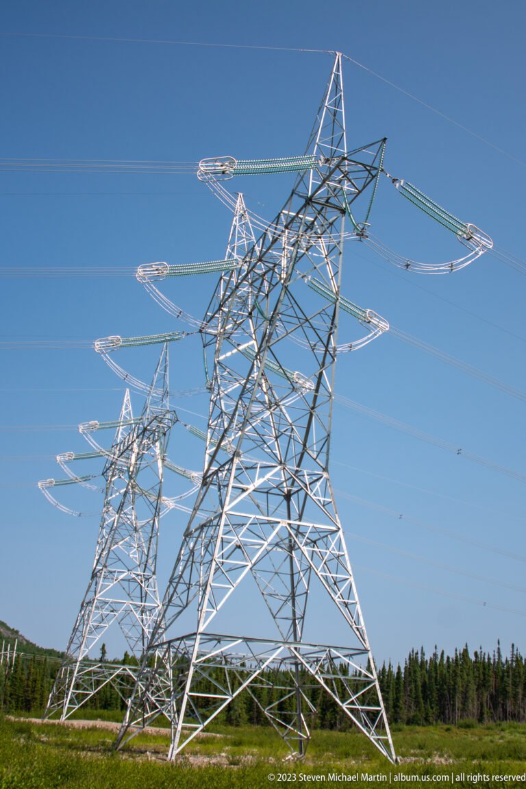 Large steel lattice transmission towers with long insulator strings and overhead conductors against a clear blue sky.