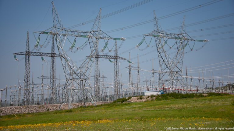 Large outdoor electrical substation with steel gantries, insulators, power lines, and transmission towers beyond a grassy foreground.