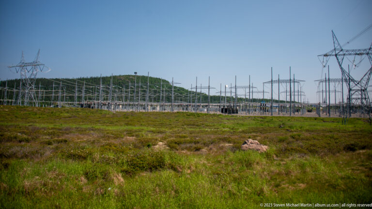 Wide electrical switchyard and transmission towers extending behind a broad foreground of low green vegetation.