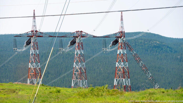 Three red-and-white lattice power towers with suspended conductors in front of a forested ridge.