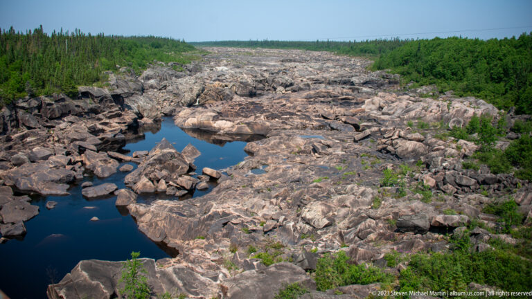 Broad exposed bedrock river channel with dark residual pools and low green vegetation along the edges.