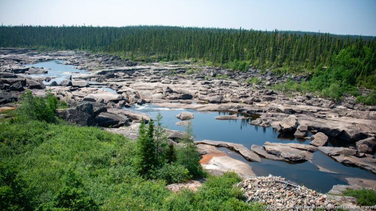 Wide exposed bedrock river channel with elongated pools and boulders, backed by dense conifer forest.