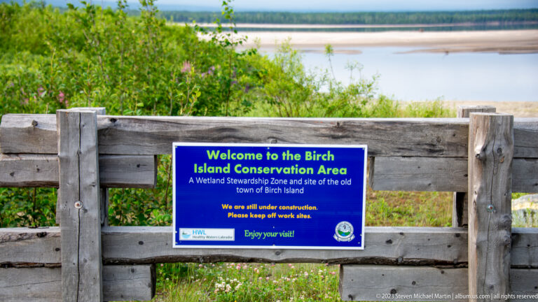 Blue Birch Island Conservation Area welcome sign on a wooden fence with wetland and open water beyond.