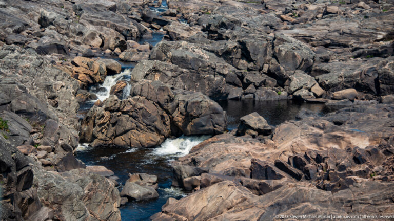 Small white cascades threading through dark fractured bedrock and pools in a mostly exposed river channel.