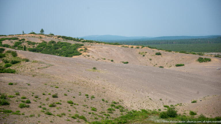 Broad sandy and gravelly open terrain with scattered shrubs, distant forest, and hazy low hills.