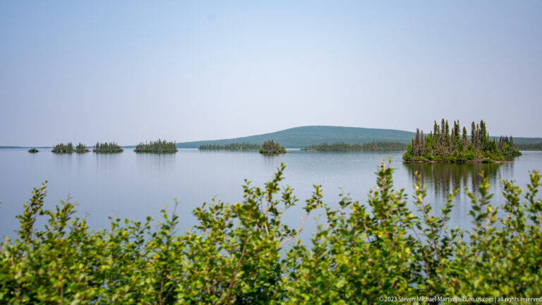 Calm lake with several small conifer-covered islands, foreground shrubs, and a low rounded hill in the distance.