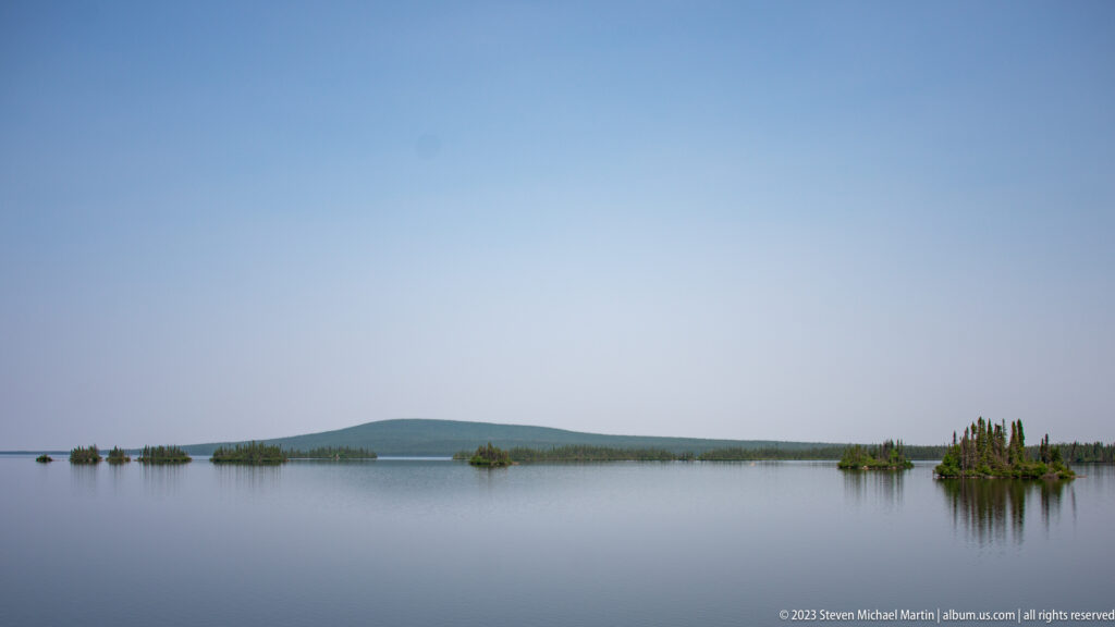 Wide calm lake beneath blue sky, with scattered wooded islets and a low hill along the distant shore.
