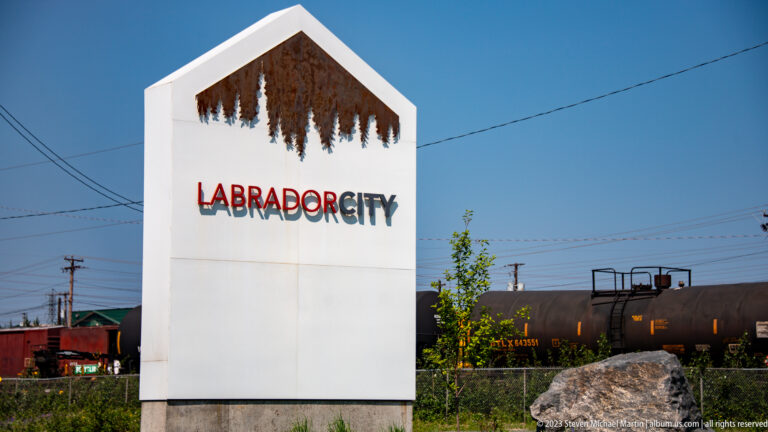 Labrador City entrance monument beside utility lines and rail tank cars under a clear blue sky.