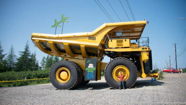 Side view of a giant yellow mining haul truck with a person standing beside one front tire for scale.