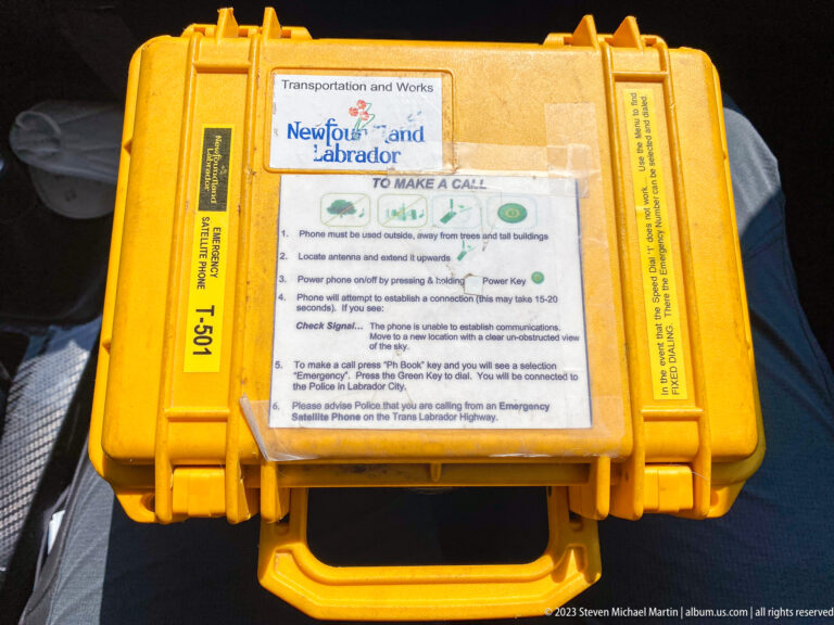 Yellow hard-shell emergency call box with Newfoundland and Labrador Transportation and Works instructions mounted on top.