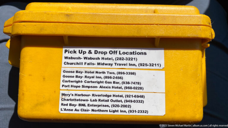 Printed pickup and drop-off location list attached to a yellow case, naming several Labrador communities and inns.