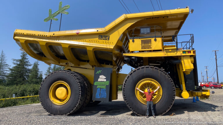 Low-angle side profile of a giant yellow mining haul truck with a person standing before one wheel.