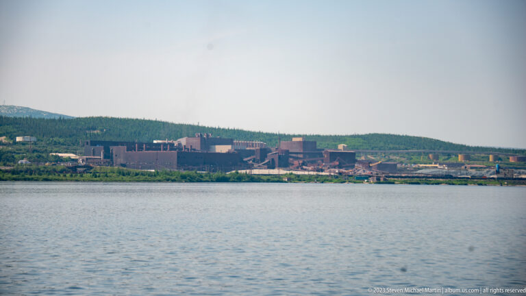 Large mining and processing complex on a distant shoreline across calm water, backed by a forested ridge.