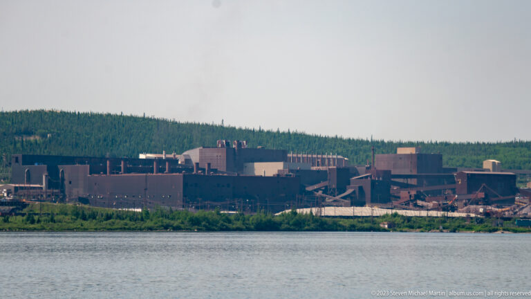 Closer view of a mining processing plant with conveyors and large block-like buildings beside water.