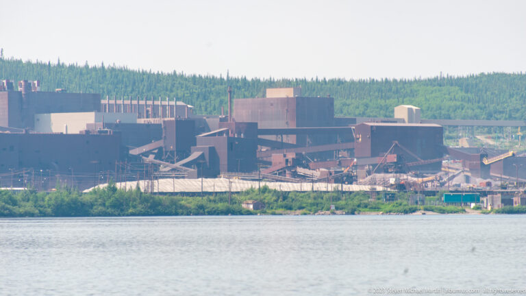 Telephoto view of large mine buildings and intersecting conveyors concentrated along a distant shoreline.