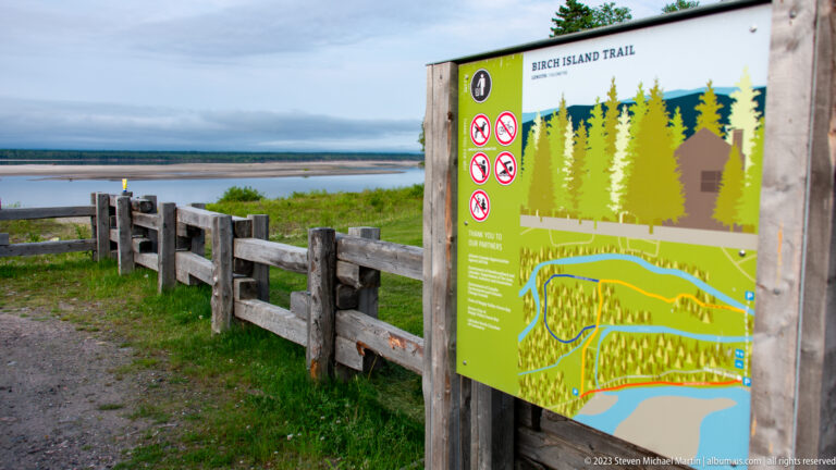 Birch Island Trail map beside a timber fence, with gravel path, wetland vegetation, and open water in the background.