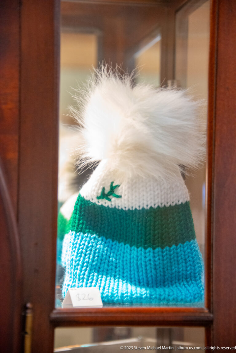 Green, turquoise, and white knitted winter hat with a large white pom-pom displayed behind glass.