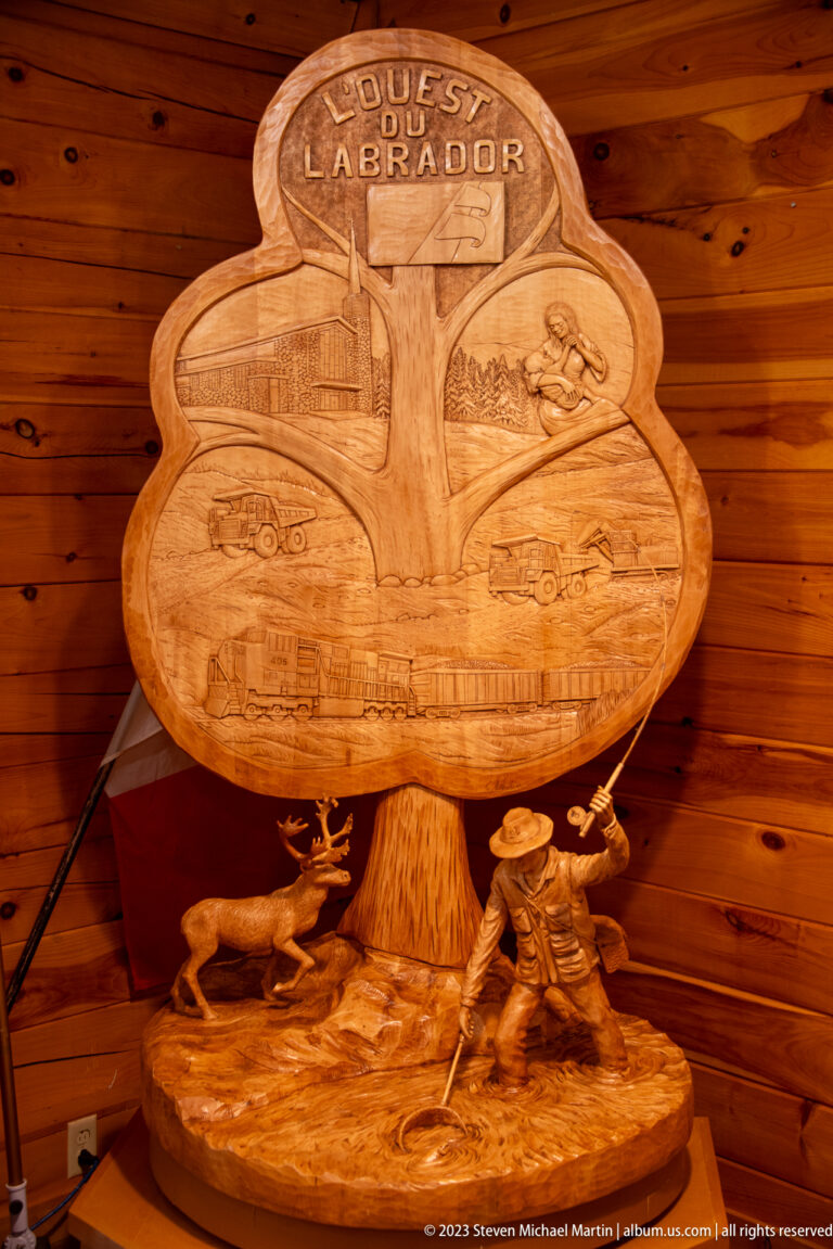 Large carved wooden relief titled “COASTS OF LABRADOR,” mounted above sculpted figures in a wood-paneled interior.