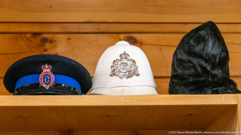 Three formal hats on a wooden shelf: a dark peaked cap, a white badged helmet, and a tall black fur-style hat.