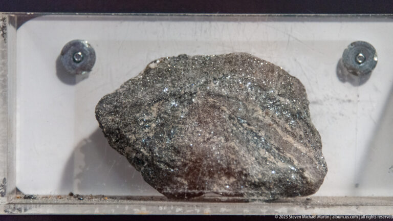Dark metallic mineral specimen with glittering surfaces displayed behind glass.