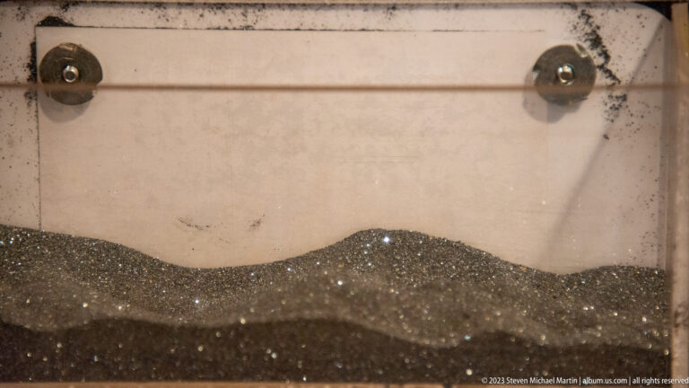 Fine dark metallic granules forming a low mound inside a clear display container.