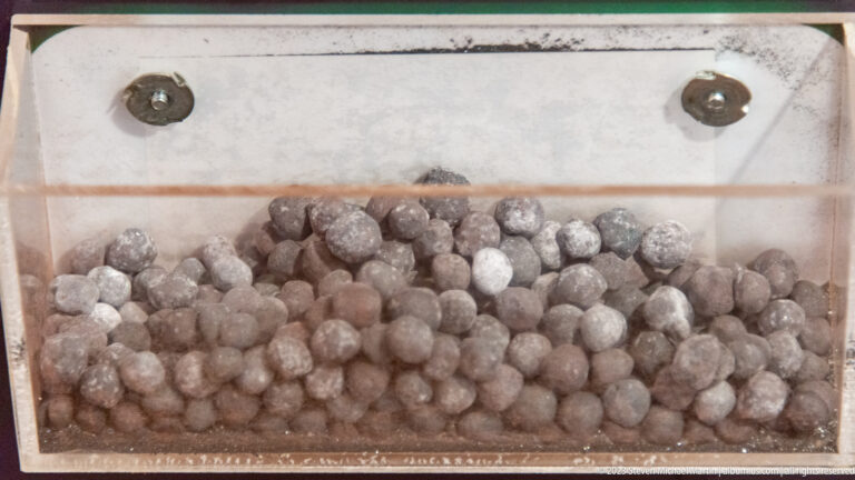 Pile of small rounded gray iron-ore pellets displayed inside a clear container.