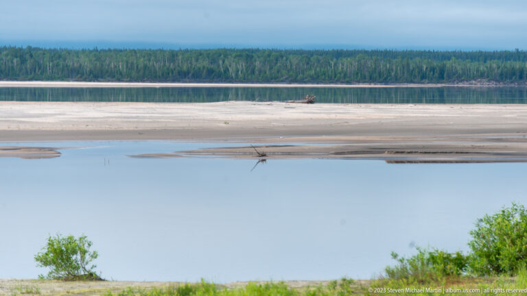 Calm water, pale exposed sandbars, and a distant forested shoreline under a cloudy sky near Birch Island.