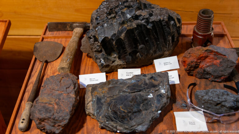 Tabletop display of large dark ore specimens, hand tools, and an industrial drill component with small labels.